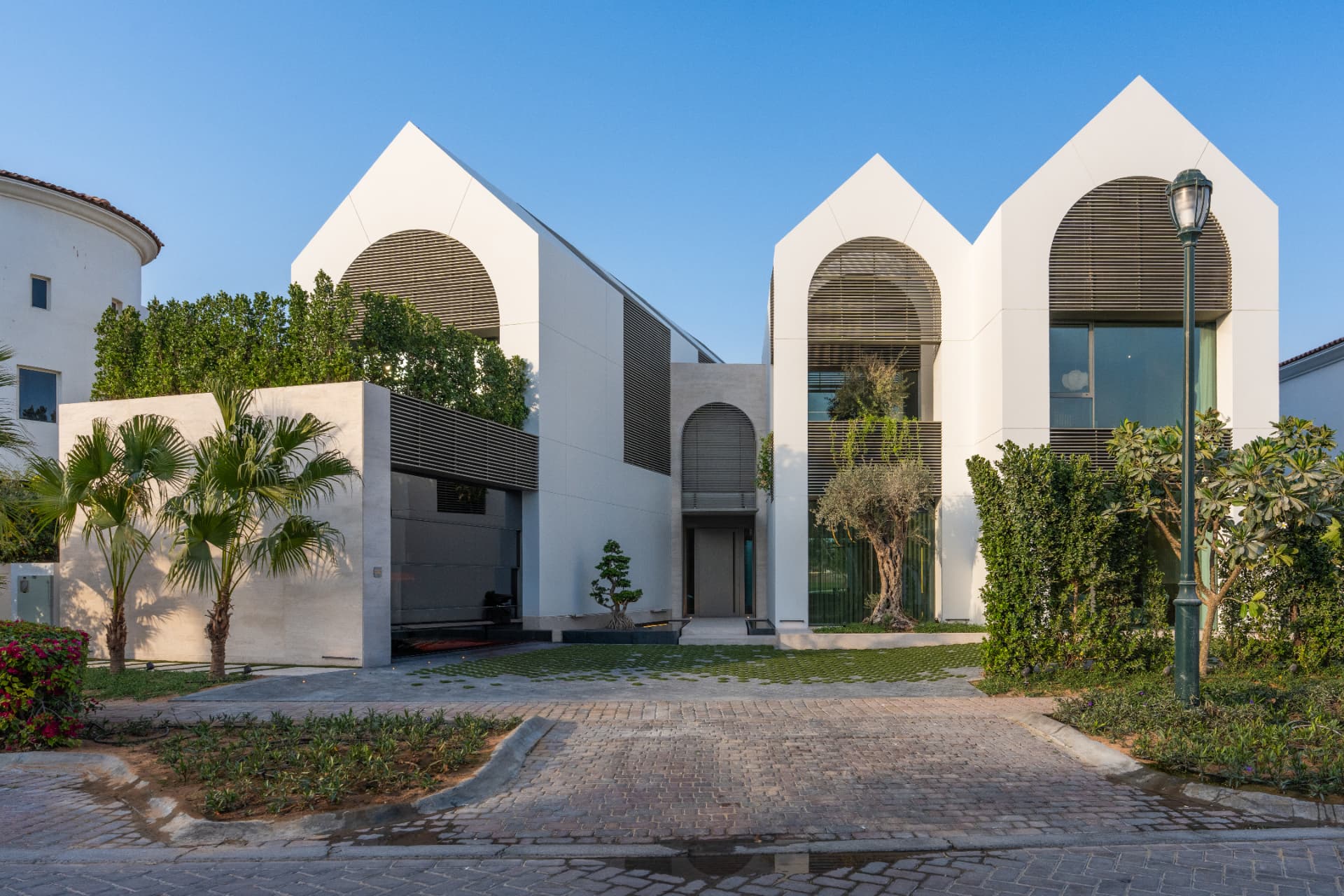 K82 street elevation by day — three white gable volumes with arched louvred screens, palms and a grass-jointed forecourt