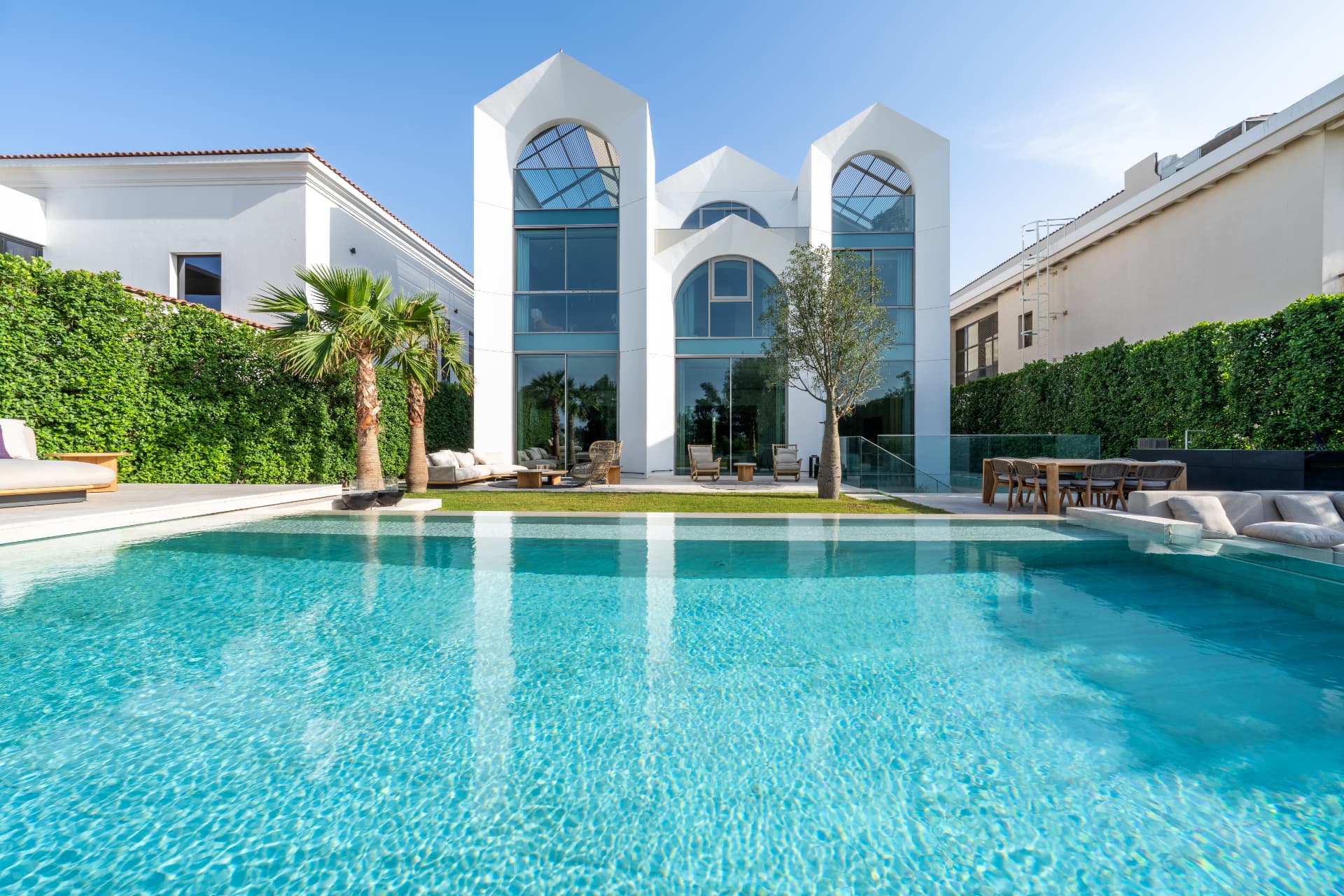 K47 rear elevation by day seen across the pool — white gabled volumes with tall arched glazing and palms