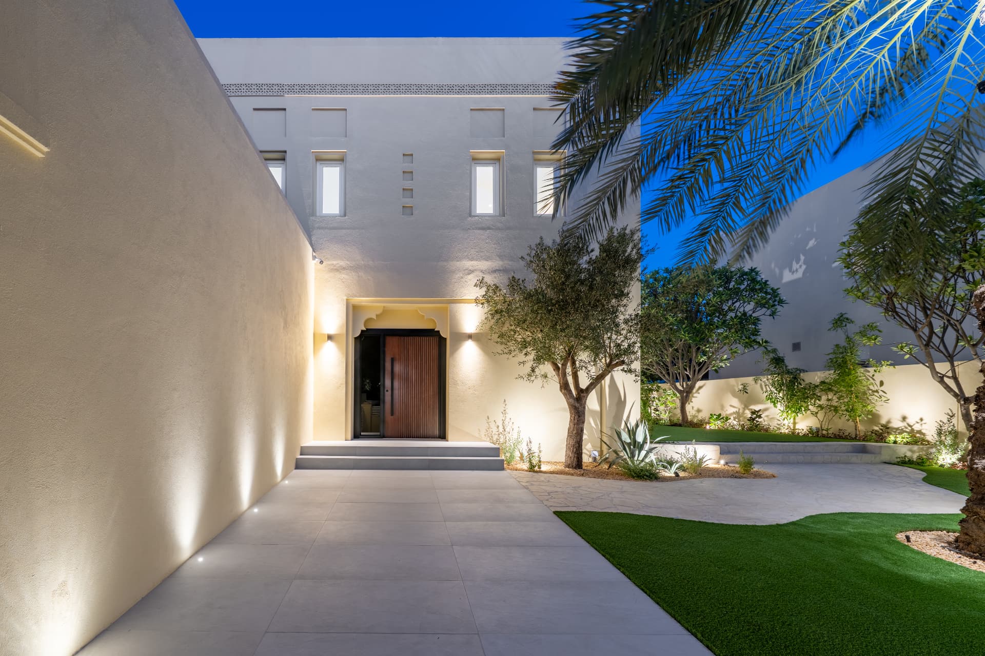 Villa 55 — verlichte entreepad met houten voordeur, olijfbomen en gazon bij blauw uur