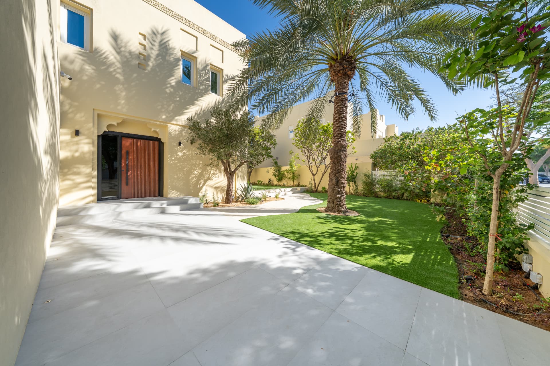 Villa 55 — voorplein overdag met houten voordeur, dadelpalm, olijfboom en gazon