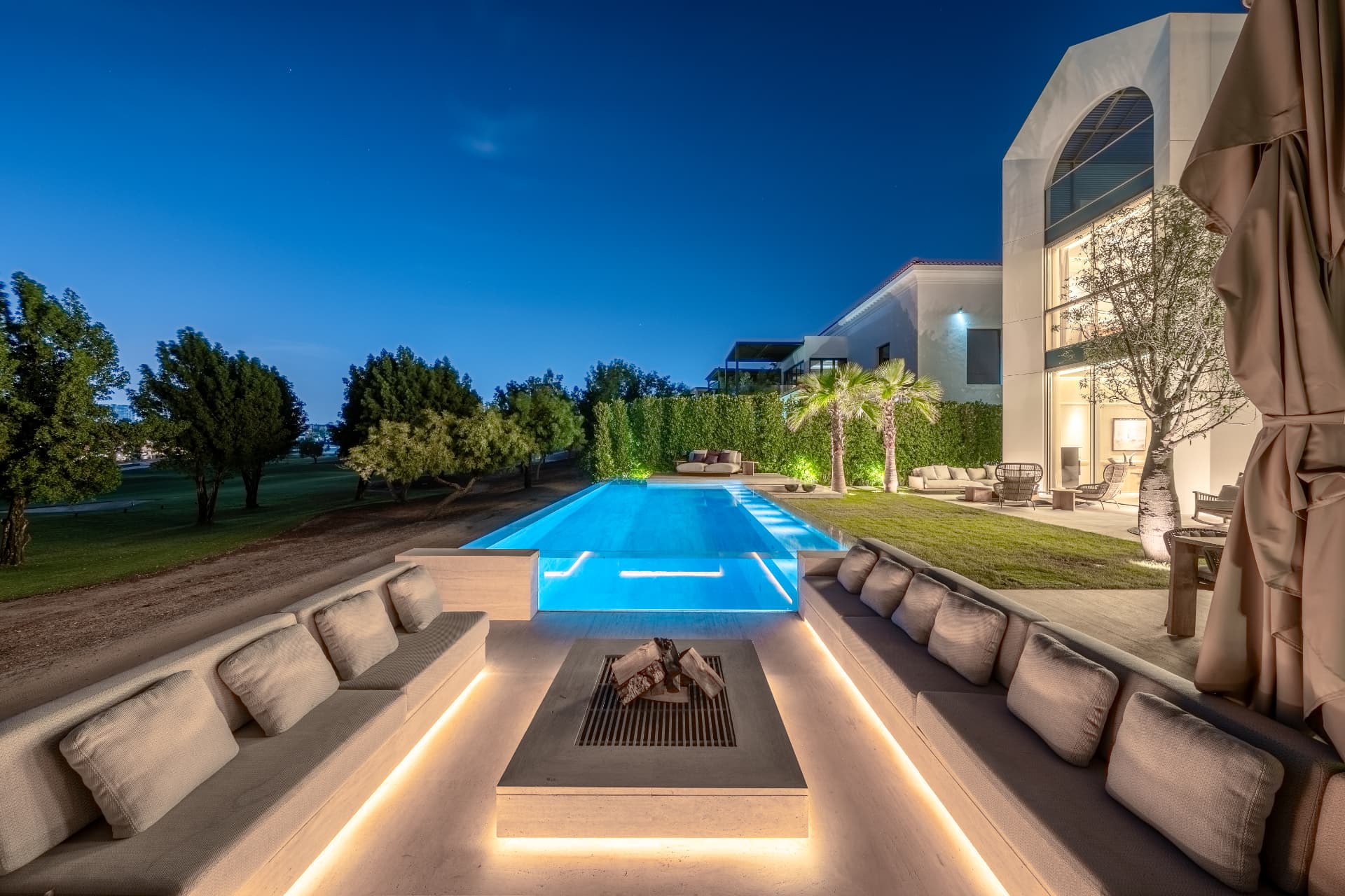 K47 sunken lounge at blue hour — travertine benches around a lit fire table beside the glass-edge lap pool