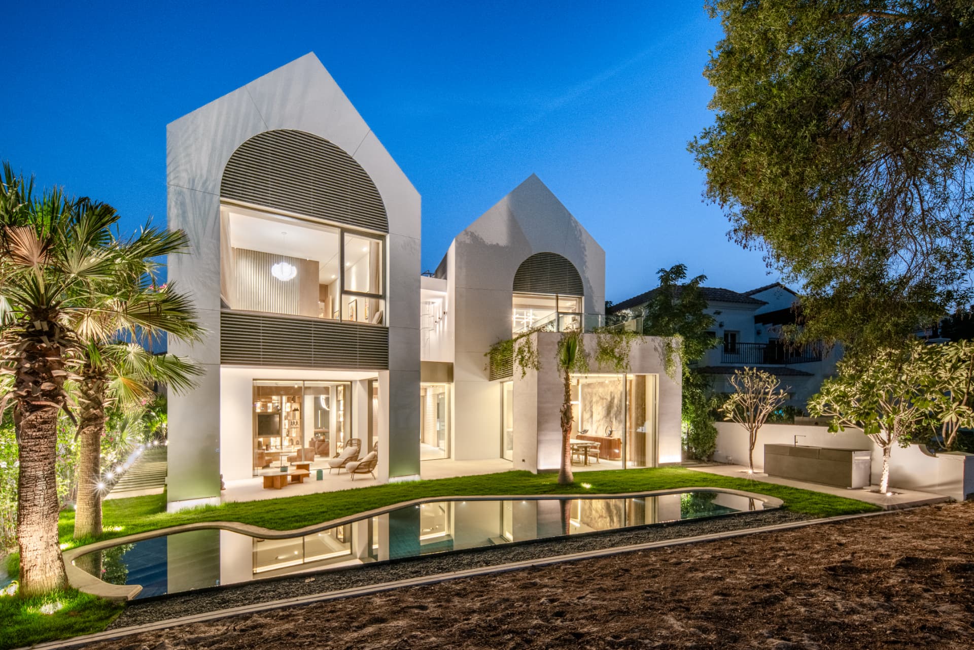 K82 rear garden at blue hour — curved pool reflecting the lit interiors of the white gable volumes