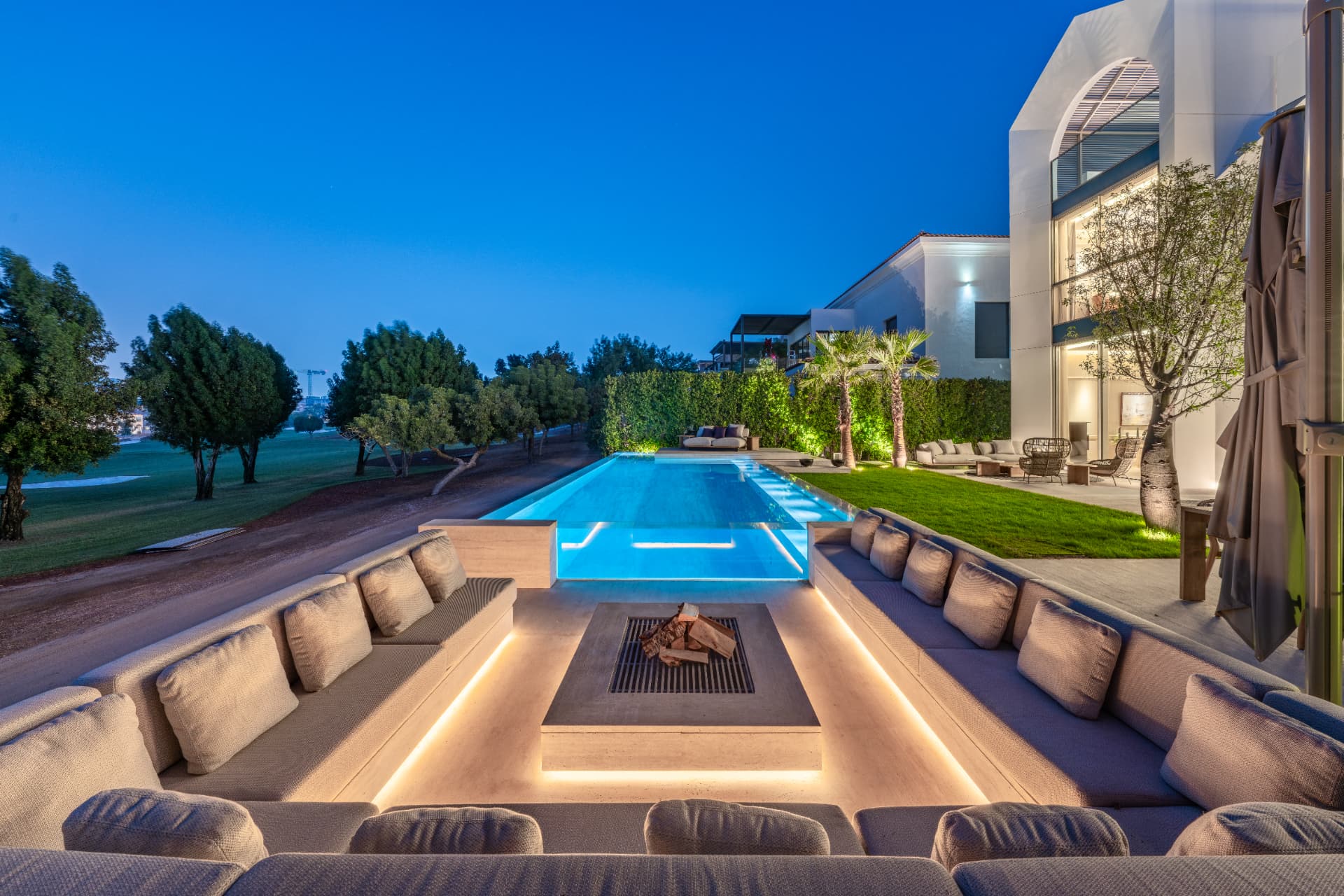 K47 sunken lounge at dusk with lit fire table, illuminated pool and uplit planting along the villa