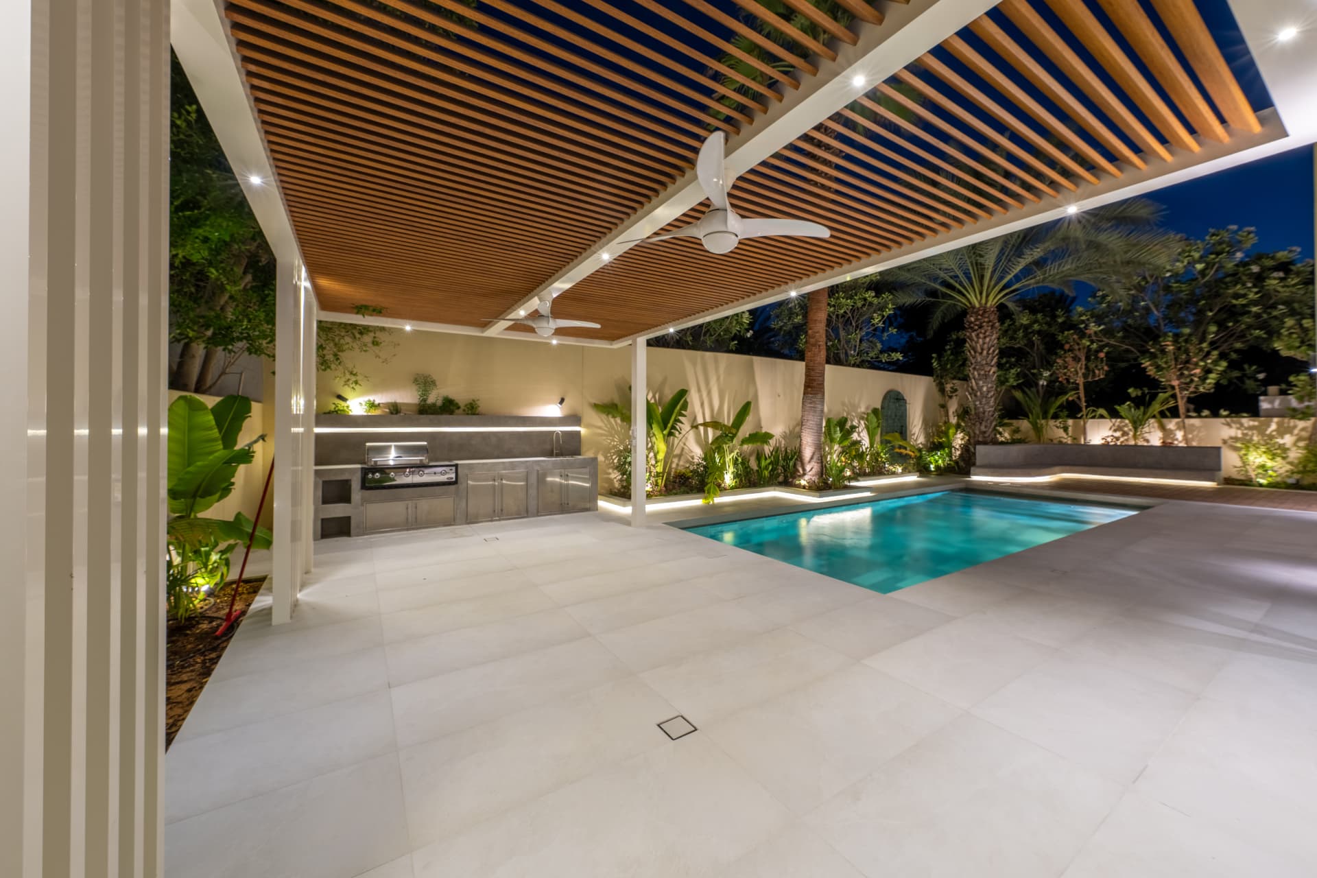Villa 55 — overdekte buitenkeuken met houten lattenplafond, ventilatoren en zwembad bij avond