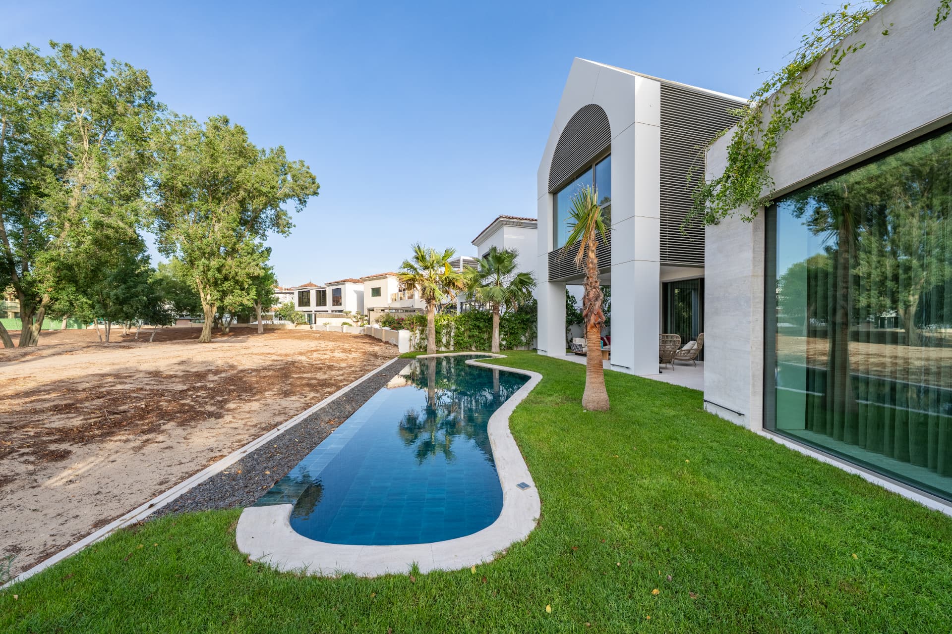 K82 — long curved pool along the lawn beside the villa with palms and full-height glazing by day