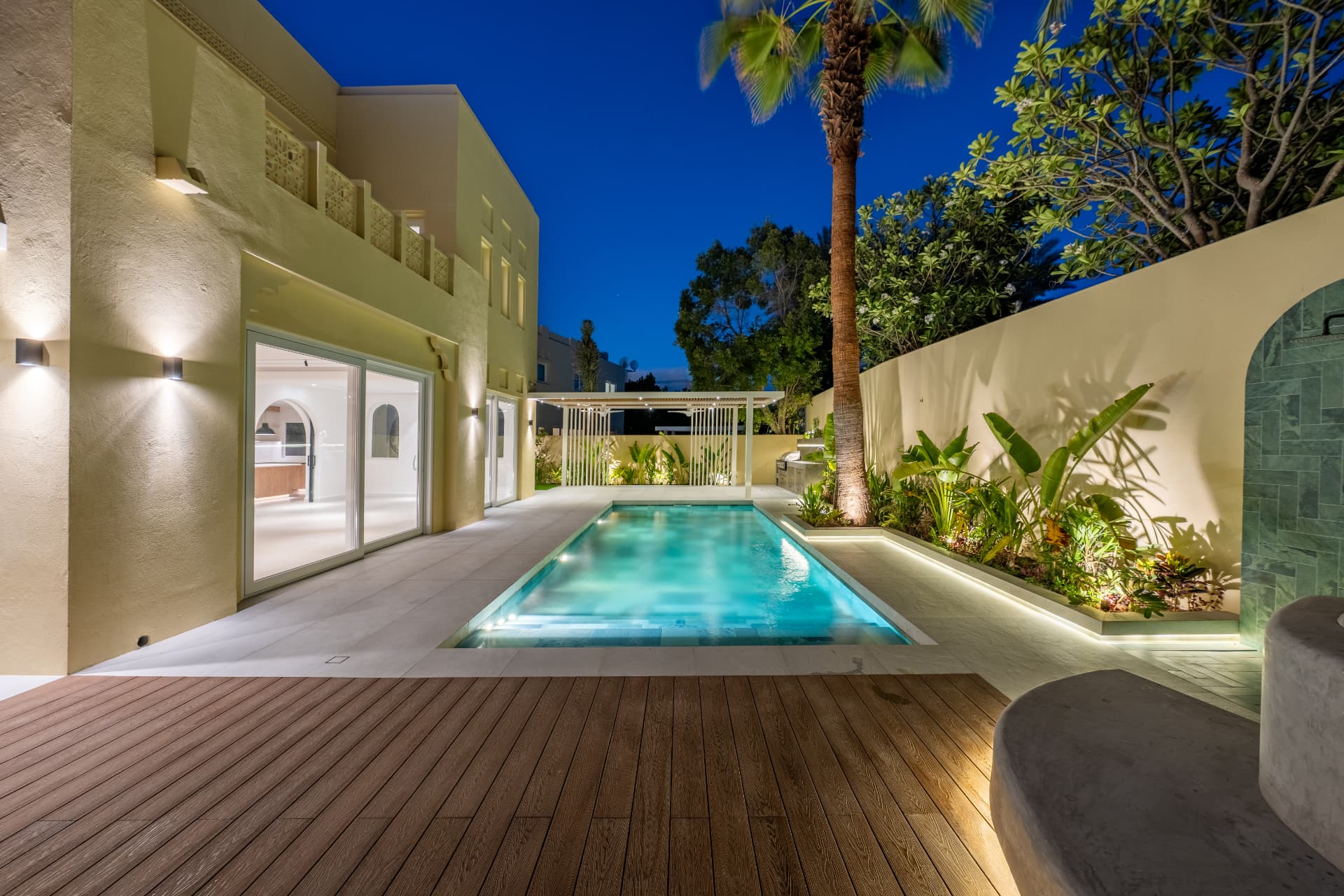 Villa 55 — verlicht zwembad met houten deck, pergola en tropische beplanting bij avond