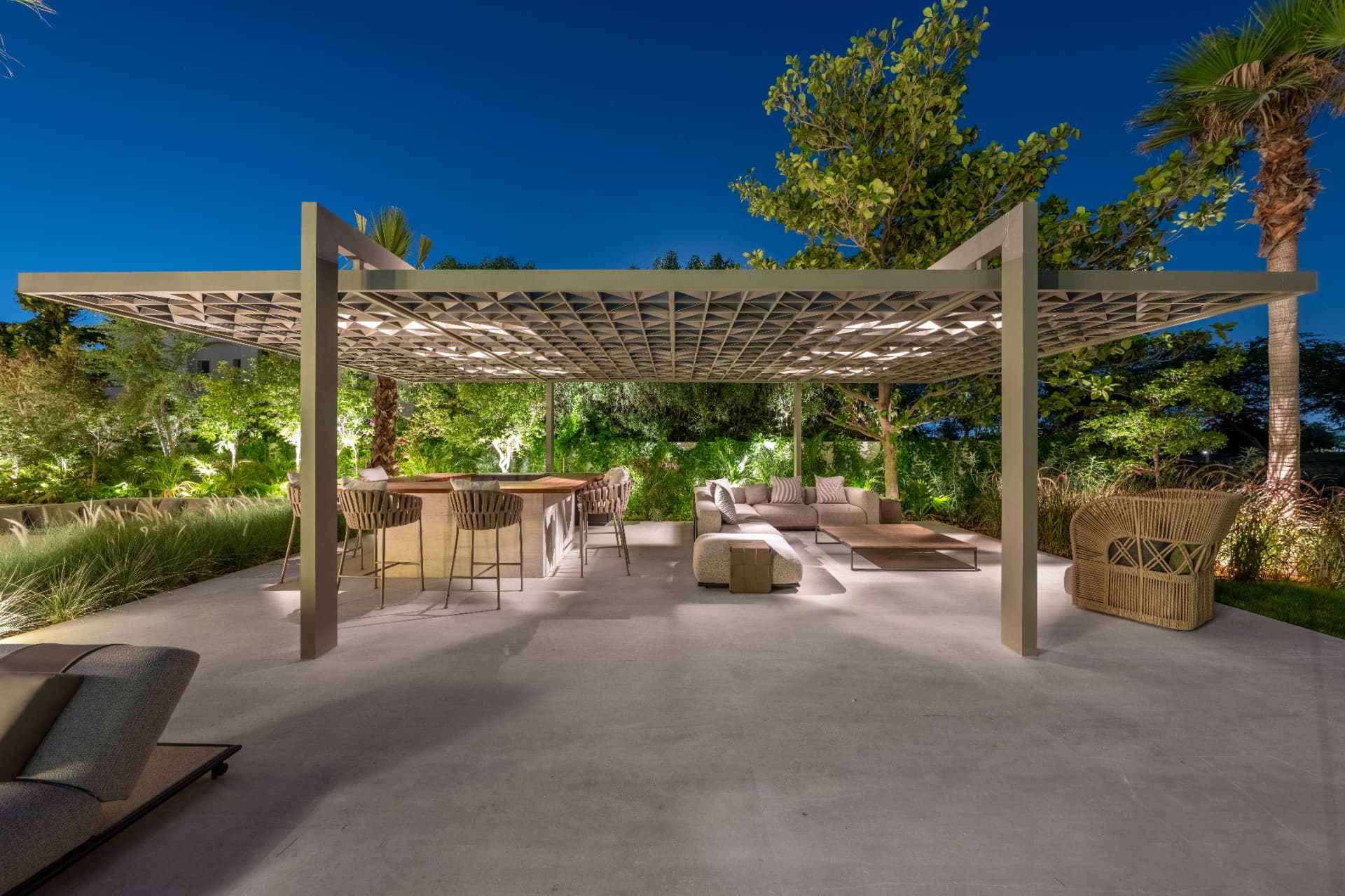 Outdoor living pavilion with latticed pergola canopy, bar seating and lounge, lit at blue hour
